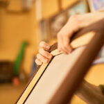 Man stretching canvas over wooden frame in art studio, hands gripping edges while working on artwork, blurred background with paintings visible